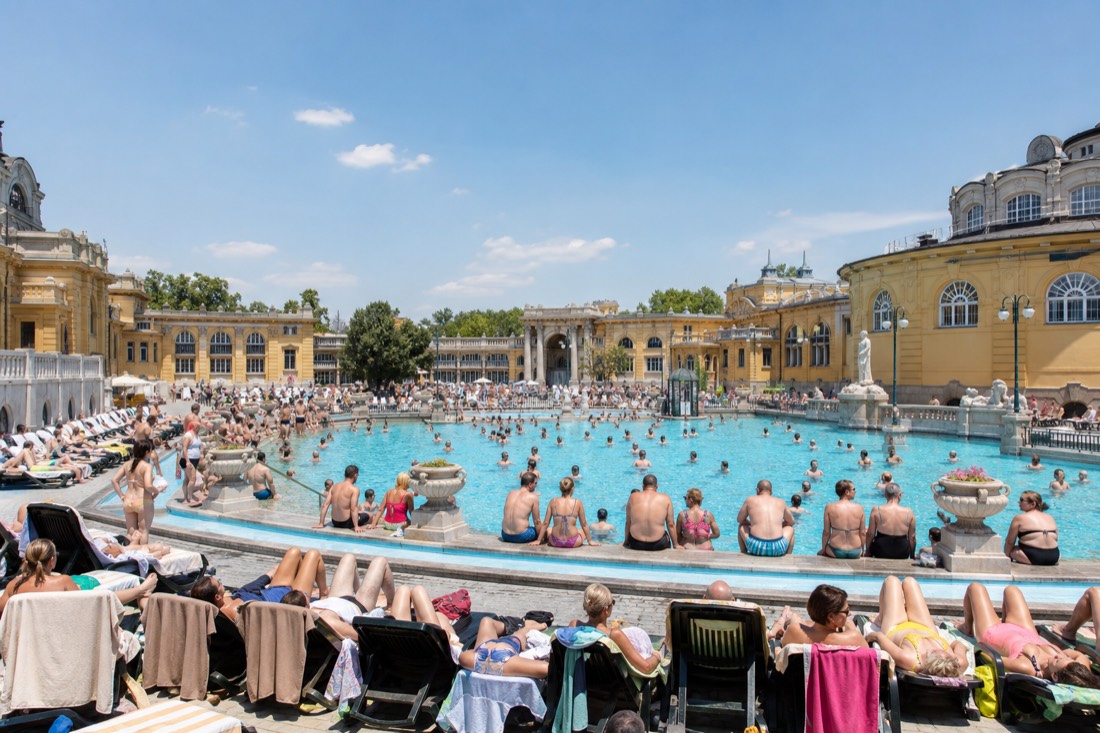 Midday crowd filling the rim of a large outdoor thermal pool at a Budapest bath while the loungers behind are all taken