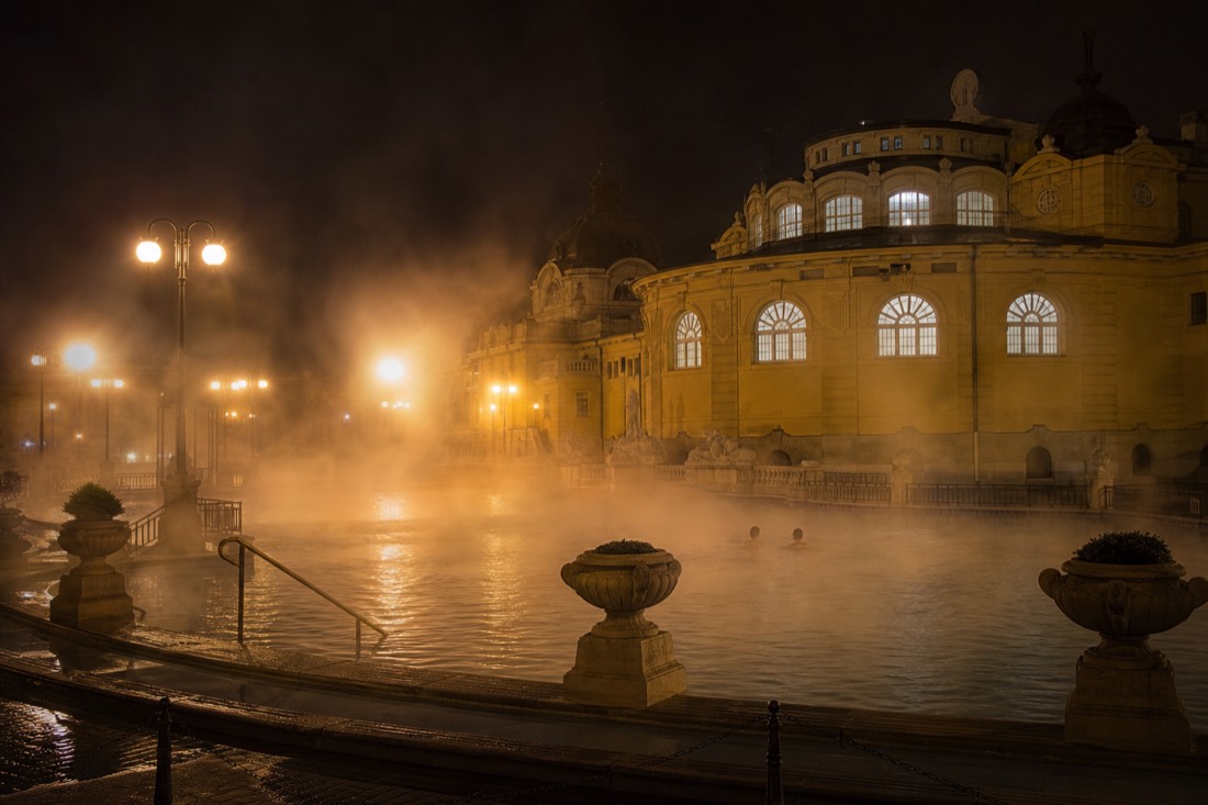 Steam glowing under warm lamps above a nearly empty outdoor thermal pool at a Budapest bath late on a Friday evening