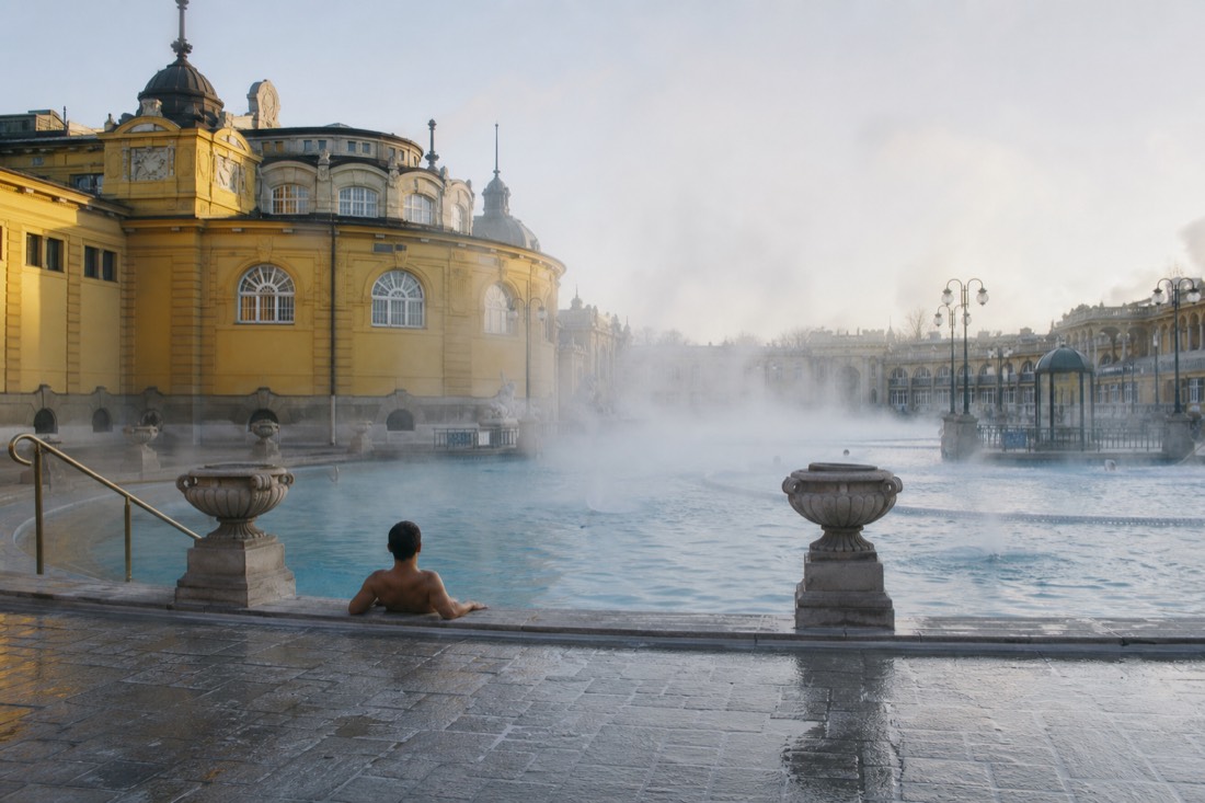 Nearly empty outdoor thermal pool at opening time with steam drifting over the water and a single bather at the edge