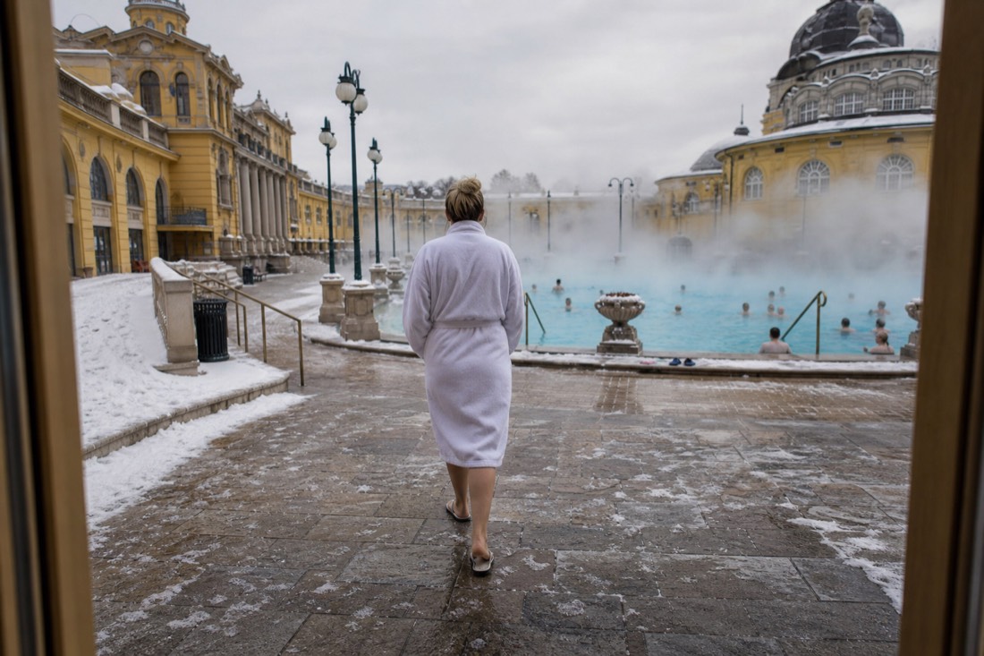 Bather in a robe walking from the door toward a steaming outdoor thermal pool across frosty stone at a Budapest bath