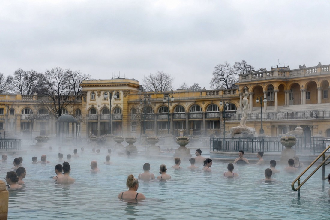 Crowded steaming outdoor thermal pool at a Budapest bath on a grey late-December afternoon with bare winter trees behind