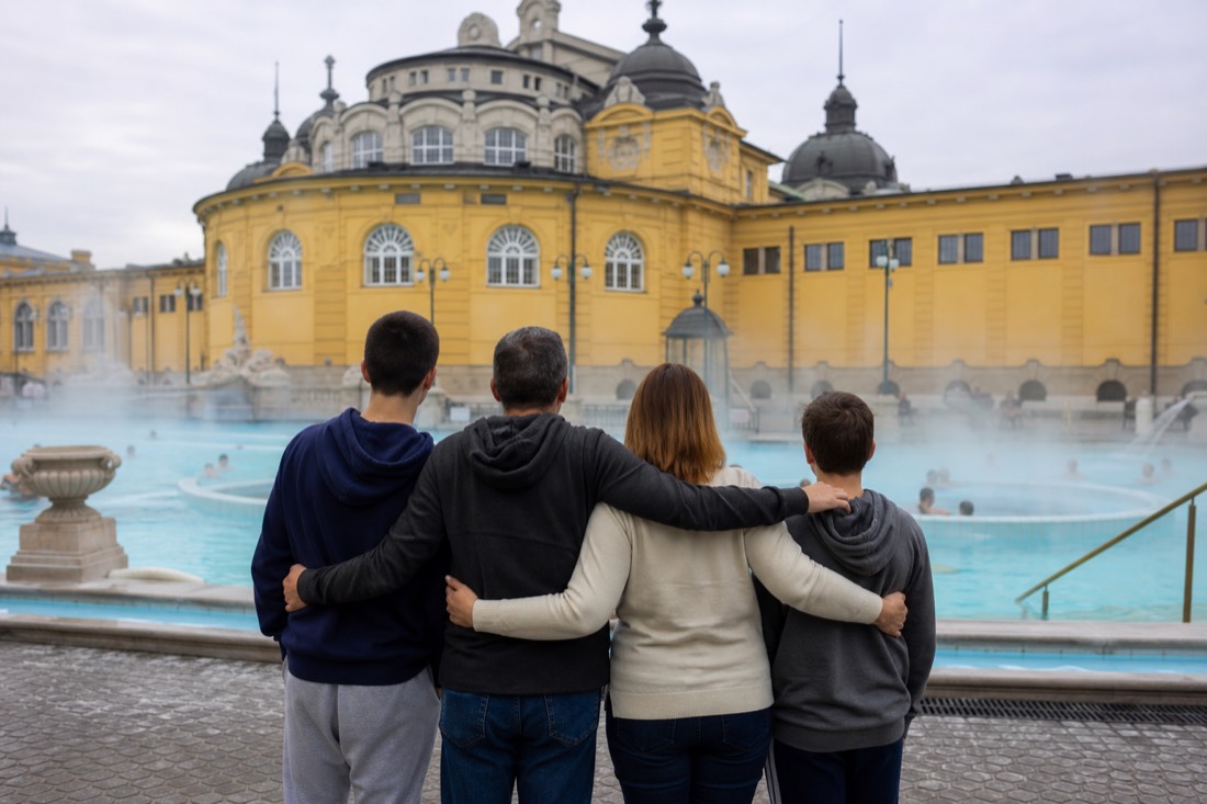 Family group of mixed ages seen from behind at the edge of a large outdoor pool in a grand Budapest bath courtyard