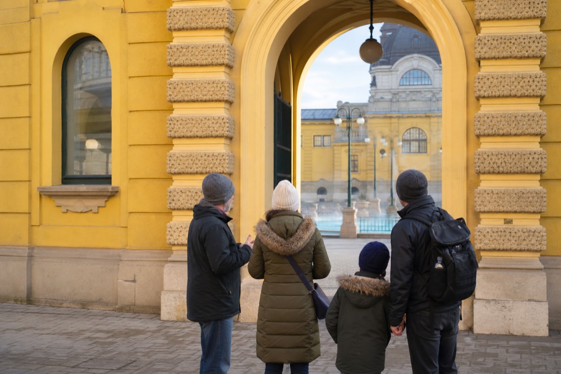 Guide talking with a family seen from behind outside the arched entrance of a yellow Budapest bath building