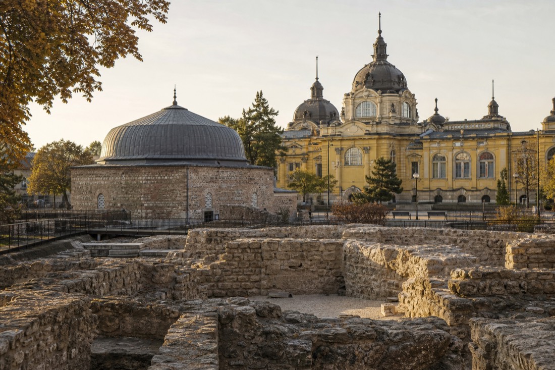 Roman stone bath foundations in the foreground with a domed Ottoman roof and a yellow palace cupola layered behind, the eras of Budapest bathing in one view