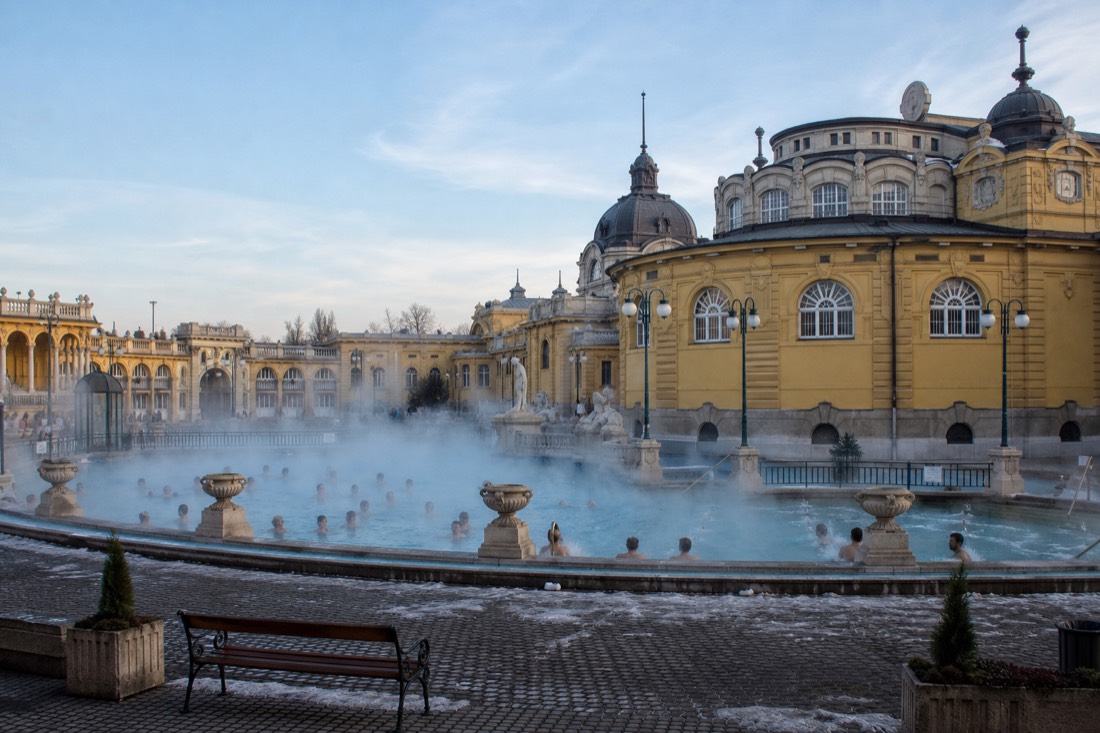 Grand neo-baroque bath palace facade in warm yellow with steam drifting over the outdoor pools