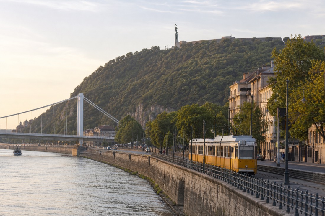 Tram running along the Buda embankment of the Danube with Gellért Hill rising behind, morning light on the river