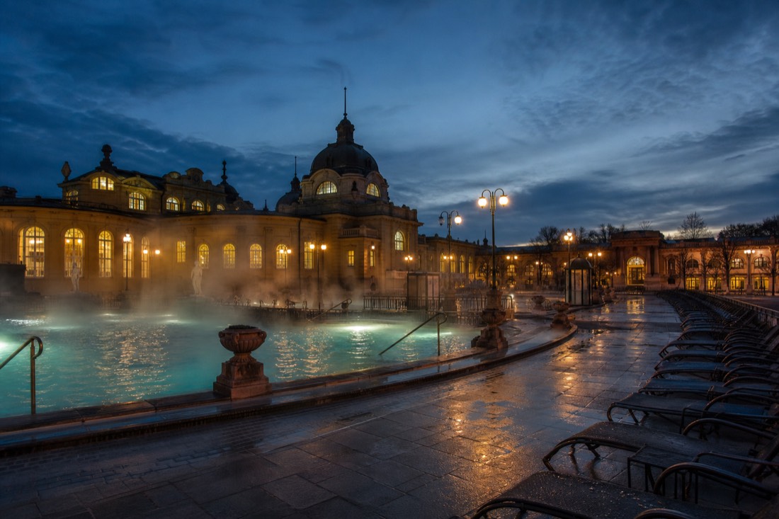 Steam drifting over an outdoor Budapest thermal pool in the evening with empty loungers