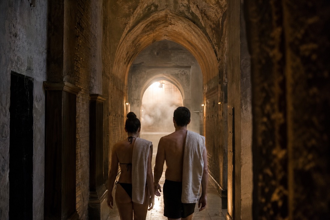 Two visitors seen from behind walking through a quiet arched passage toward warm light in a historic Buda bath