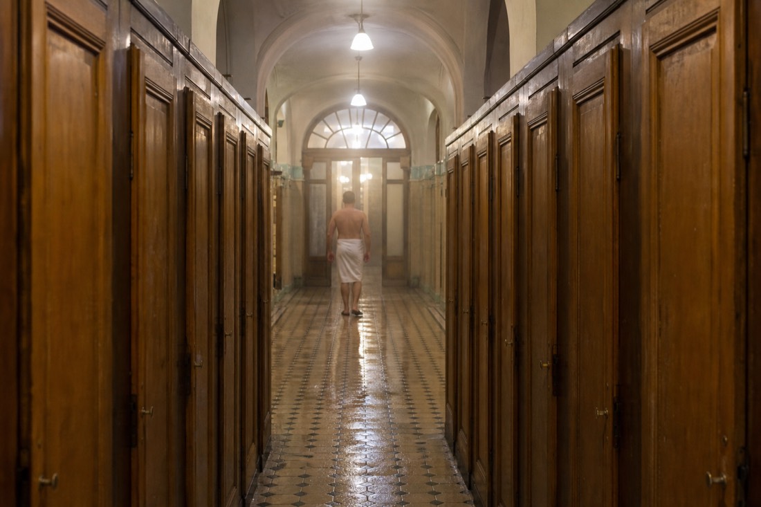 Long tiled changing-hall corridor in a historic Budapest bathhouse with plain wooden cabin doors and a wet floor
