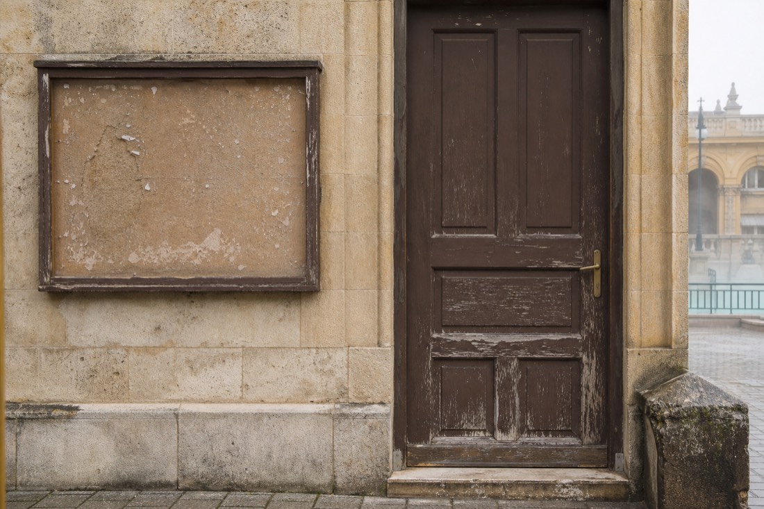 Weathered notice board beside the closed door of a small historic Budapest bathhouse